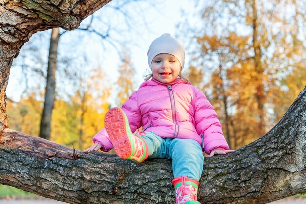 Glückliches junges Mädchen, das auf Baum im schönen Herbstpark auf Naturspaziergängen im Freien lächelt und sitzt. Kleines Kind, das im orange gelben Hintergrund des Herbstes spielt. Hallo Herbstkonzept.