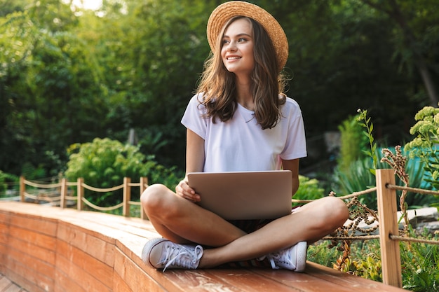 Glückliches junges Mädchen, das mit Laptop-Computer am Park draußen sitzt