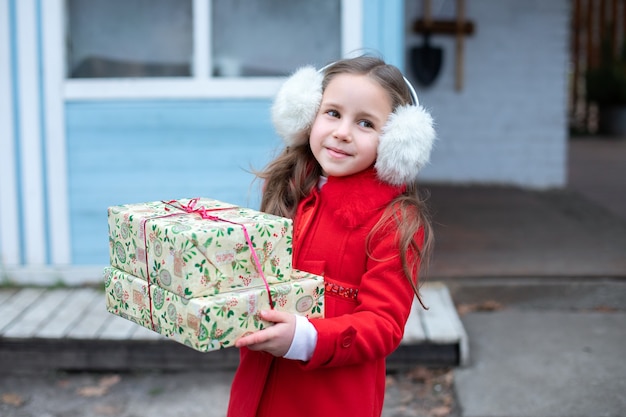 Glückliches junges Mädchen im roten Mantel, das an der Weihnachtsstraße mit Geschenken steht