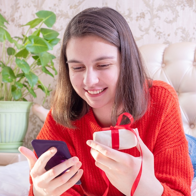 Glückliches junges Mädchen in einem roten Pullover, der ein Geschenk hält und Familie oder Freunde mit einem Smartphone, Nahaufnahme, Vorderansicht, Quadrat gratuliert.