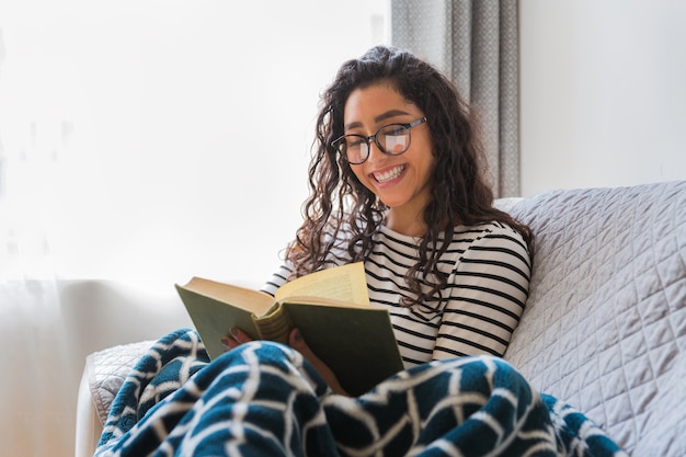 Foto glückliches junges mädchen liest ein buch in ihrem wohnzimmer. sie hat eine decke an den beinen