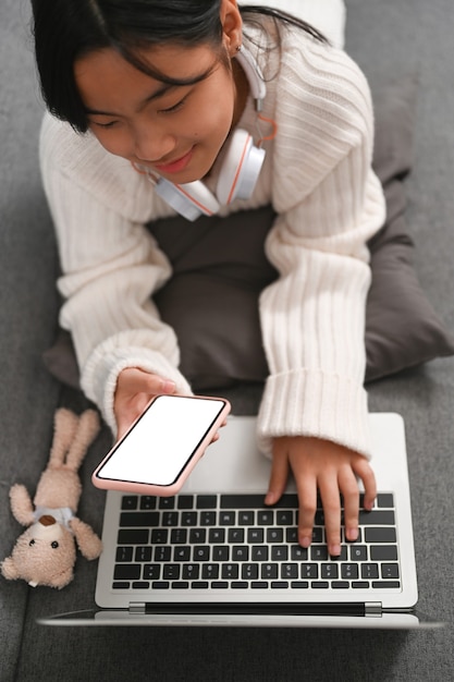 Glückliches junges Mädchen mit Smartphone und Laptop beim Liegen auf dem Sofa zu Hause.