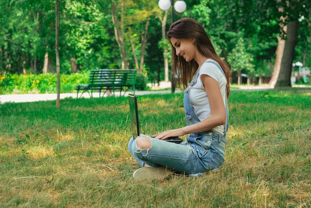 Glückliches junges Mädchen sitzt im Sommer auf dem Gras und schaut auf Laptop