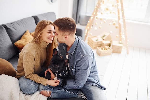 Glückliches junges modernes Paar, das zu Hause mit Weihnachtsbaum mit ihrem süßen Hund auf dem Sofa sitzt.