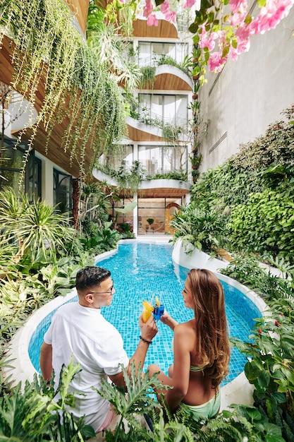 Glückliches junges Paar, das am Pool sitzt und mit leckeren Cocktails anstößt?