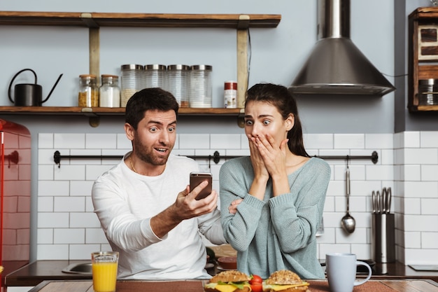 Glückliches junges Paar, das an der Küche während des Frühstücks zu Hause sitzt, mit Handy