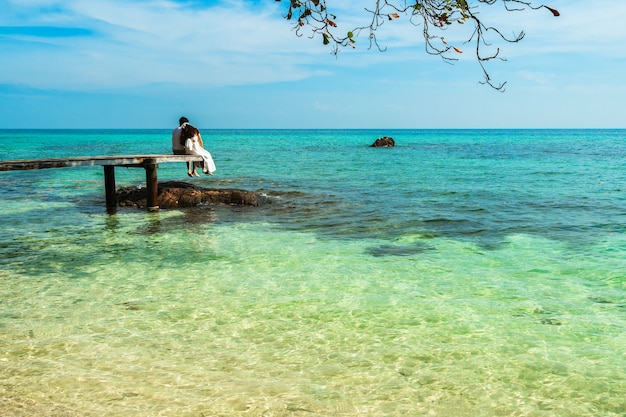 Glückliches junges Paar, das auf einer Holzbrücke und einem Meeresstrand auf Koh MunNork Island, Rayong, Thailand sitzt