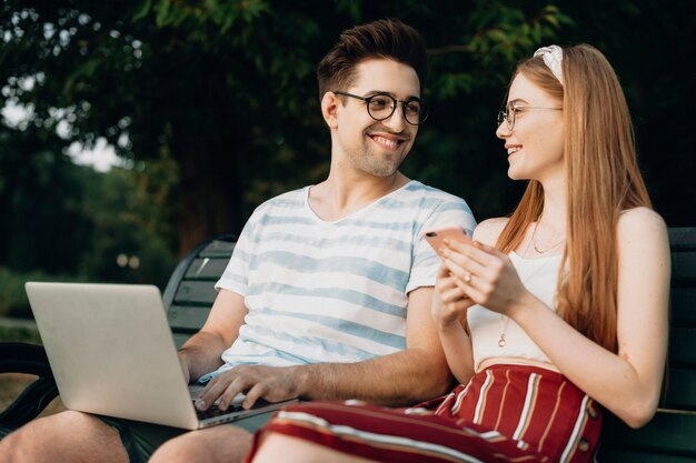 Glückliches junges Paar, das beim Sitzen auf einer Bank im Park arbeitet. Junger Programmierer, der Datencode schreibt, während er mit seiner Freundin sitzt.