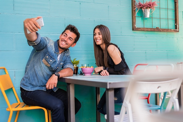 Glückliches junges Paar, das in einem Restaurant mit einem Smartphone sitzt, das ein Selfie nimmt