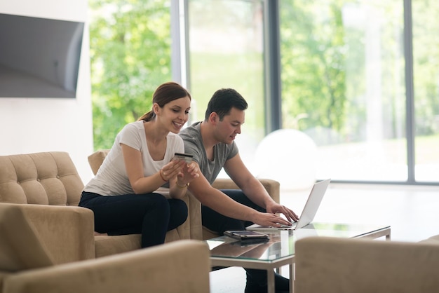 glückliches junges Paar, das online mit einem Laptop, einem Computer und einer Kreditkarte in seiner luxuriösen Villa kauft