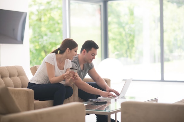 glückliches junges Paar, das online mit einem Laptop, einem Computer und einer Kreditkarte in seiner luxuriösen Villa kauft