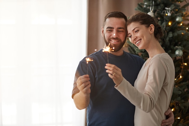 Glückliches junges Paar, das sich gegen den Weihnachtsbaum umarmt und brennende Wunderkerzen betrachtet