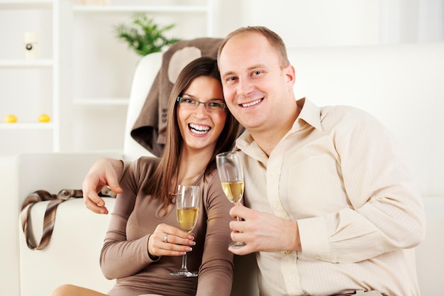 Glückliches junges Paar, das zu Hause umarmt sitzt, lächelt und Champagner trinkt.