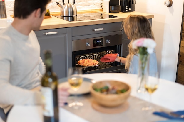 glückliches junges Paar, das zu Hause zu Abend isst, und eine junge schöne blonde Frau, die eine Pizza aus dem Ofen nimmt