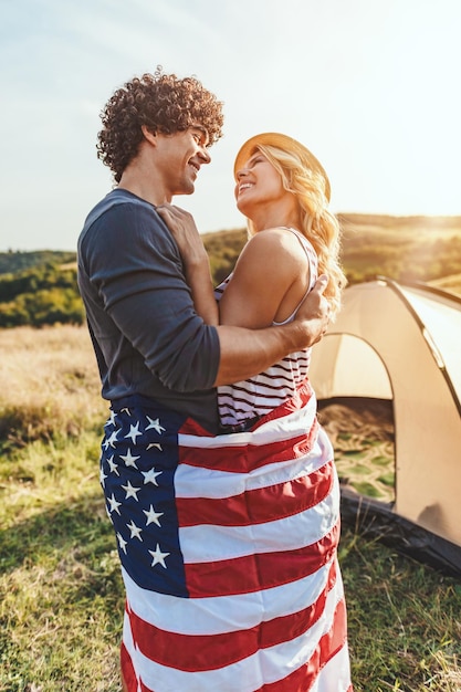 Glückliches junges Paar genießt einen sonnigen Tag in der Natur. Sie umarmen sich, eingehüllt in amerikanische Flaggen, vor einem Campingplatzzelt.