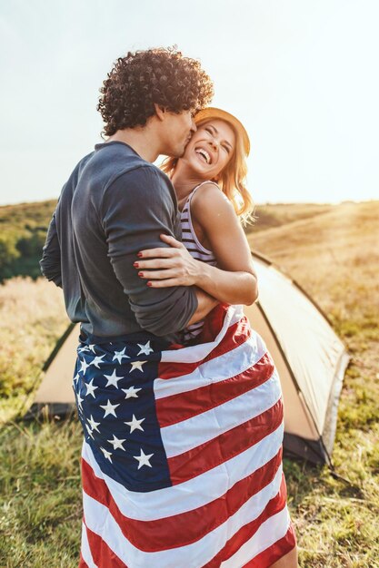 Glückliches junges Paar genießt einen sonnigen Tag in der Natur. Sie umarmen sich, eingehüllt in amerikanische Flaggen, vor einem Campingplatzzelt.