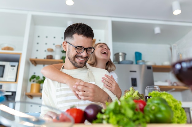 Glückliches junges Paar hat Spaß in der modernen Küche drinnen und bereitet frischen Obst- und Gemüsesalat zu Schönes junges Paar spricht und lächelt, während es zu Hause gesundes Essen in der Küche kocht