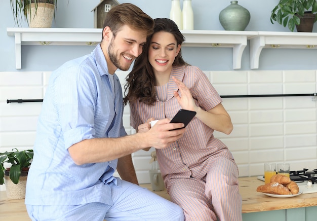 Glückliches junges Paar im Pyjama, das Online-Inhalte auf einem Smartphone ansieht und beim Kochen in der Küche zu Hause lächelt.