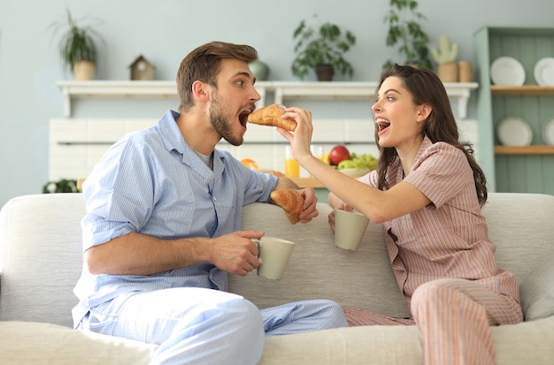 Glückliches junges Paar im Pyjama in der Küche beim Frühstück, einander ein Croissant füttern.