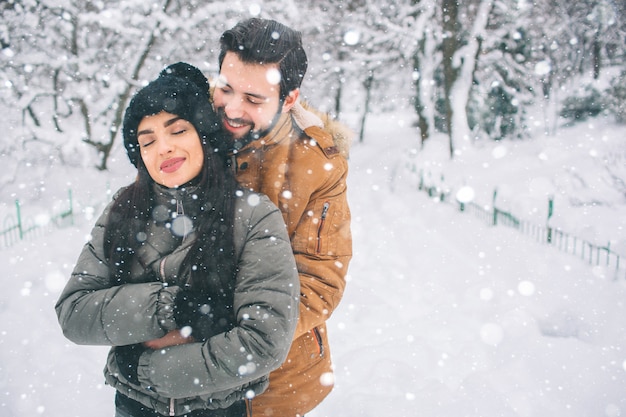 Glückliches junges Paar im Winter. Familie draußen. Mann und Frau, die aufwärts schauen und lachen. Liebe, Spaß, Jahreszeit und Leute - gehend in Winterpark. Es schneit, sie umarmen sich