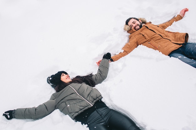 Glückliches junges Paar im Winter. Familie draußen. Mann und Frau, die aufwärts schauen und lachen. Liebe, Spaß, Jahreszeit und Leute - gehend in Winterpark. Im Neuschnee liegen, Schneeengel zum Spaß bringen