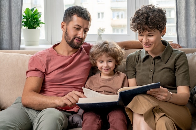 Glückliches junges Paar und ihr süßer kleiner Sohn, der durch Seiten der großen Enzyklopädie schaut, während sie auf bequemer Couch durch Fenster sitzen