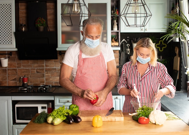 Glückliches kaukasisches gealtertes Paar, das zu Hause in der Gesichtsmaske kocht. Salat zu Hause machen. Hausmannskost. Soziale Distanz. Kochen während der Covid 19.