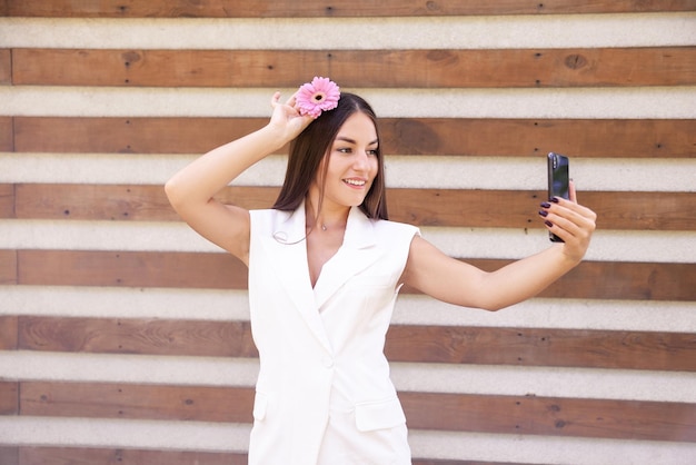 Glückliches kaukasisches Mädchen in weißer Kleidung mit einer rosa Blume im Haar, das Selfies am Telefon macht