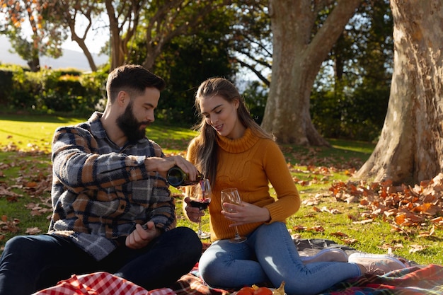 Glückliches kaukasisches Paar sitzt auf einem Teppich im sonnigen Herbstgarten und trinkt Wein. Häusliches Leben, Freizeit, Romantik und Zweisamkeitskonzept.