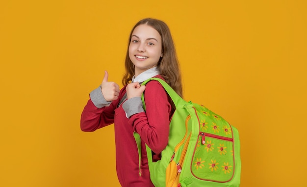 Glückliches Kind, das am 1. September Daumen mit Schultasche auf gelbem Hintergrund gibt