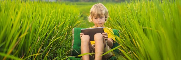 Glückliches kind, das auf dem feld sitzt und einen tablet-jungen hält, der an einem sonnigen tag zu hause auf dem gras sitzt
