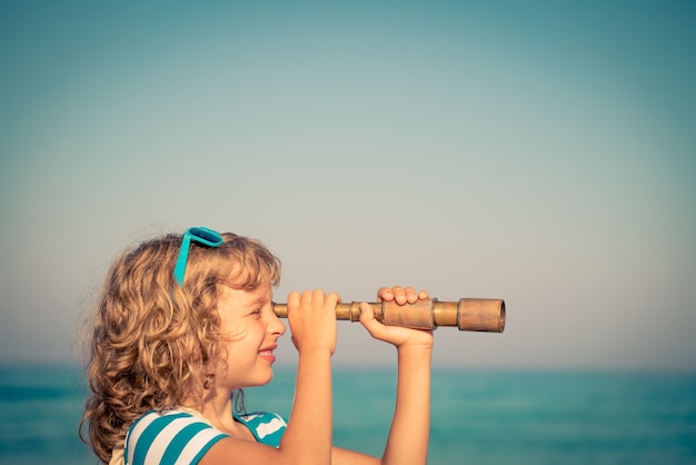 glückliches Kind, das im Sommer ein Fernglas gegen Meer- und Himmelshintergrund verwendet