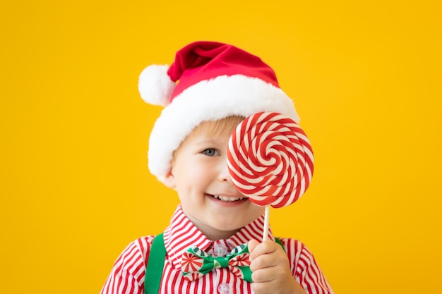 Glückliches Kind, das in der Hand Lutscher gegen gelben Hintergrund hält. Porträt des lustigen Kindes kleidete Weihnachtsmann-Hut an. Weihnachtsferienkonzept