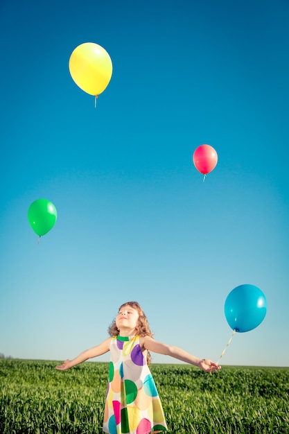 glückliches Kind, das mit hellen mehrfarbigen Ballons im Freien spielt, das Spaß im grünen Frühlingsfeld hat