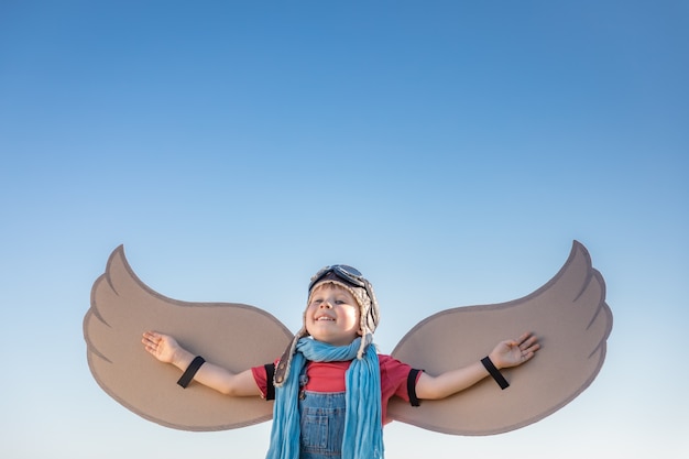 Glückliches Kind, das mit Spielzeugflügeln gegen Hintergrund des blauen Himmels spielt