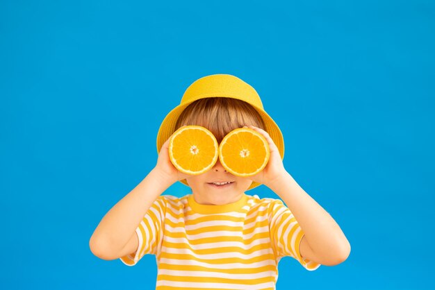 Glückliches Kind, das Scheiben von Orangenfrüchten wie Sonnenbrillen hält. Kind, das gestreiftes gelbes T-Shirt gegen blauen Papierhintergrund trägt. Konzept für gesunde Ernährung und Sommerferien