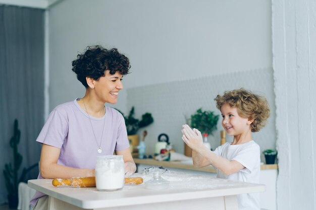 Glückliches Kind, das Spaß mit Mehlklatschen hat, das zu Hause Gebäck mit Mutter kocht