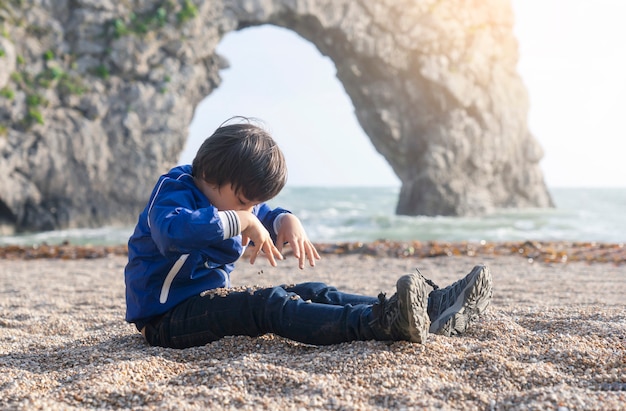 Glückliches Kind, das unten schaut, spielend mit Kieseln auf seinen Beinen durch das Meer mit undeutlichem Hintergrund