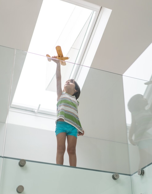 Glückliches Kind, das zu Hause das hölzerne Flugzeug steht unter dem Dachfenster träumt hält