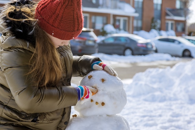 Glückliches Kind der Weihnachtszeit, das mit einem Schneemann im verschneiten Winter spielt ein junges Mädchen formt Schneemann