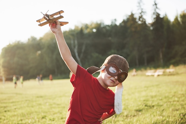 Glückliches Kind im Pilotenhelm, der mit einem hölzernen Spielzeugflugzeug spielt und davon träumt, fliegen zu werden