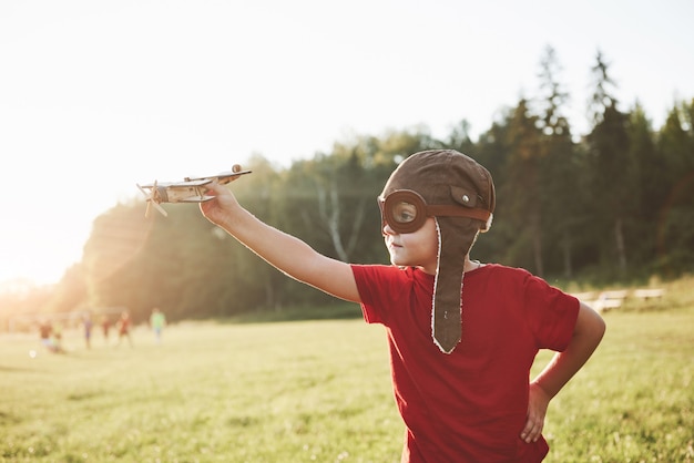 Glückliches Kind im Pilotenhelm, der mit einem hölzernen Spielzeugflugzeug spielt und davon träumt, fliegen zu werden