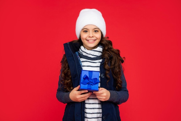 Glückliches Kind in Pufferjacke und Hut hält Box-Kind mit anwesendem Teenager-Mädchen auf rotem Hintergrund