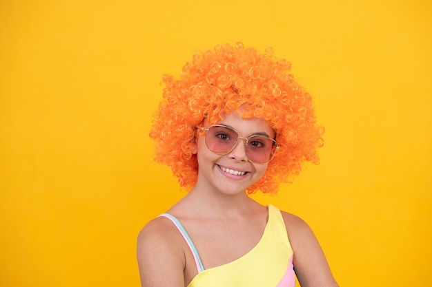 Glückliches Kind in Sonnenbrille und Badeanzug mit orangefarbenem, lockigem Perückenhaar auf gelbem Hintergrund, Sommerspaß.