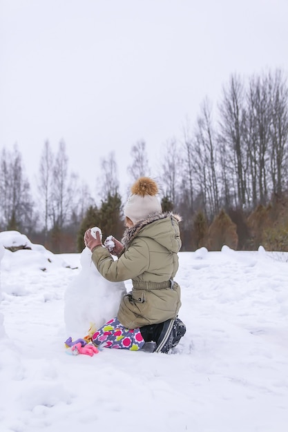 Glückliches Kind macht einen Schneemann auf einem schneebedeckten Feld auf dem Land.