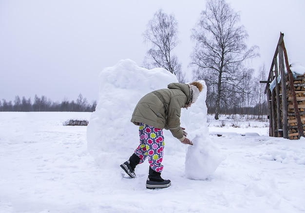 Glückliches Kind macht einen Schneemann auf einem schneebedeckten Feld auf dem Land.