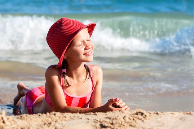 Glückliches Kind Mädchen in Rot liegt am Sonnenstrand auf Meereswellenhintergrund Lächelnder Teenager, der das Meer genießt