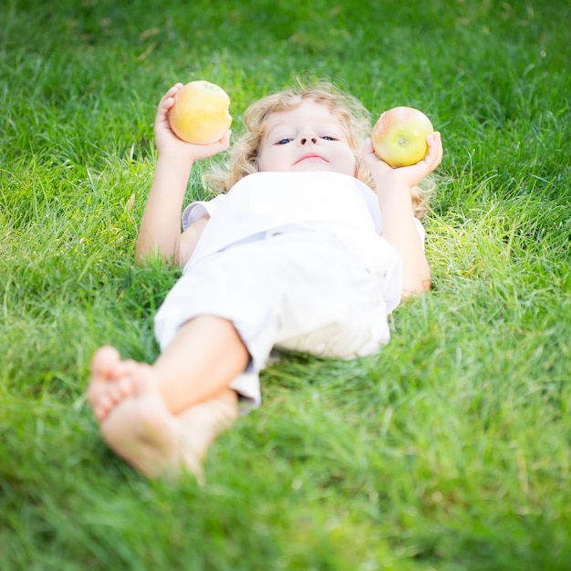 Glückliches Kind mit Äpfeln, die im Frühlingspark auf grünem Gras liegen