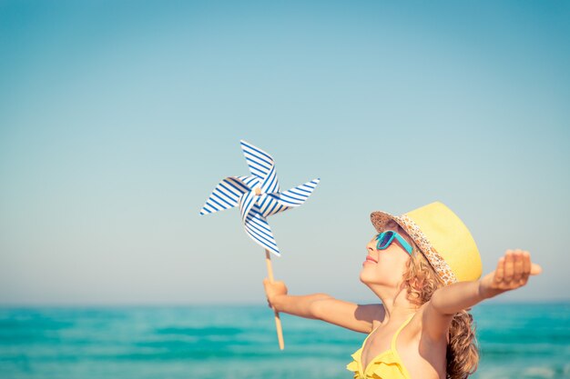 glückliches Kind mit Sonnenbrille und Hut, Windrad und offenen Händen vor blauem Meer- und Himmelshintergrund