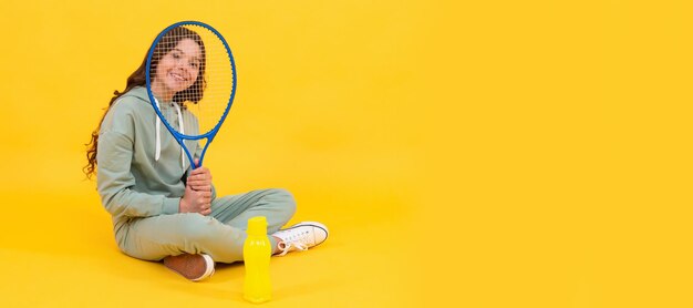 Glückliches Kind sitzt in Sportbekleidung mit Squashschläger und Wasserflasche auf gelbem Hintergrund Horizontal